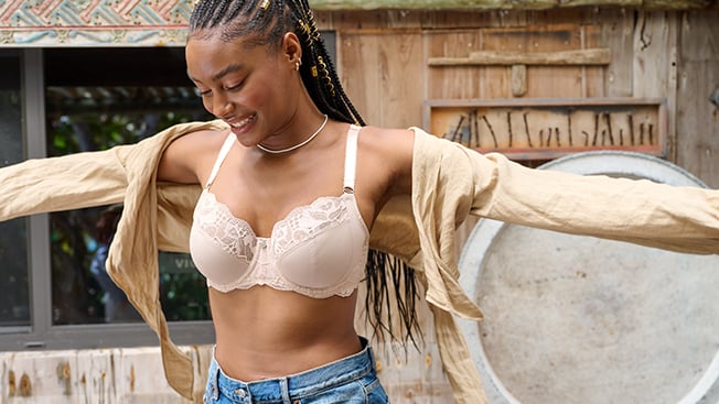 Woman with her arms wide outdoors in frnt of a wooden building, with her shirt open to reveal a white lacey Fantasie bra.